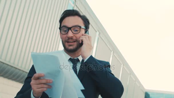 Belgeler, belgeler tutan cep telefonuyla konuşurken çekici şık bir iş adamı. Adam konuşmayı. Yakın çekim. Açık havada şehirde ofis binası yakınındaki — Stok video