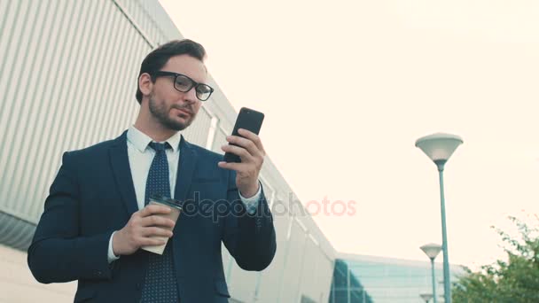 Empresário atraente usando smartphone, navegar na internet, mensagens de texto e beber café fora do prédio do centro de escritórios. Fechar — Vídeo de Stock