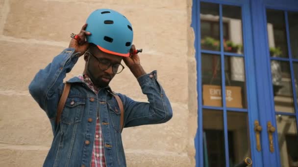 Retrato de un joven afroamericano en ropa casual con gafas y bicicleta usando casco y sonriendo en la cámara . — Vídeo de stock