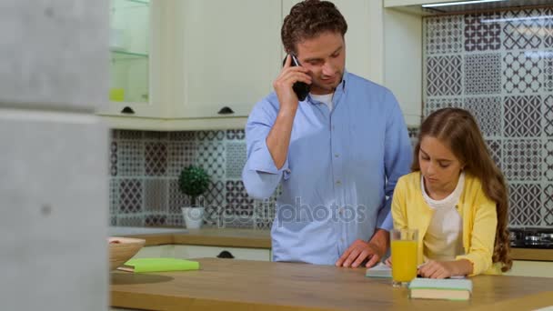 Nette Tochter macht Hausaufgaben in der Küche, während ihr Vater mit dem Smartphone telefoniert. — Stockvideo