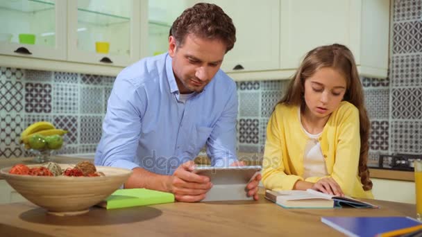 Schattig jong meisje het lezen van het boek bij de keuken terwijl haar vader met behulp van Tablet PC. — Stockvideo