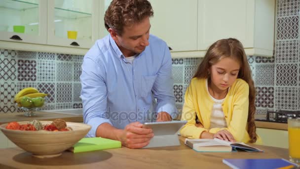 Atractivo padre joven ayudando a su hija con su tarea en la cocina usando tableta y hablando con ella . — Vídeo de stock
