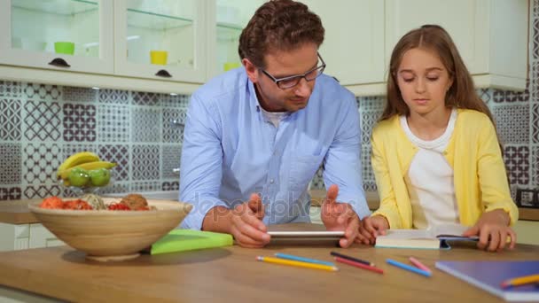 Young father in the glasses using tablet, his daughter reading the book at the kitchen than beating head of father and start laughting. — Stock Video