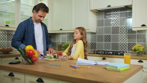 Jovem menina feliz com cabelo loiro fazendo os trabalhos de casa na cozinha, enquanto seu pai cortando o pepino para o almoço para a salada . — Vídeo de Stock