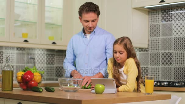 Ung familj i köket, far skära sallad medan dotter använder surfplatta för att titta på videon. — Stockvideo