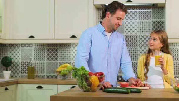 Familia caucásica en la cocina haciendo una ensalada juntos, padre cortando el tomate, hija bebiendo el jugo de naranja . — Vídeo de stock