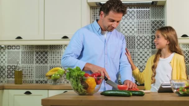 Familia caucásica en la cocina haciendo una ensalada juntos, padre cortando el tomate, hija usando tableta . — Vídeo de stock