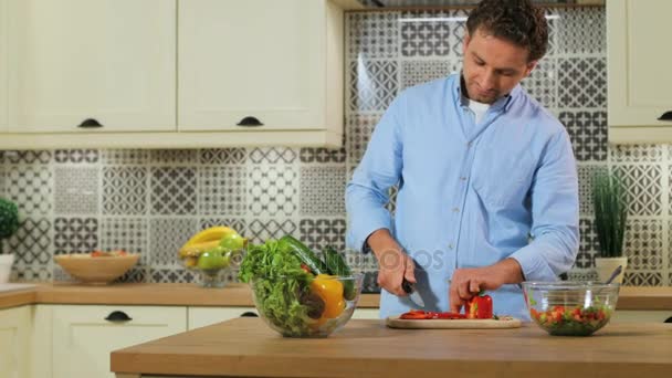 Белый мужчина режет перец для салата на деревянном столе на кухне . — стоковое видео