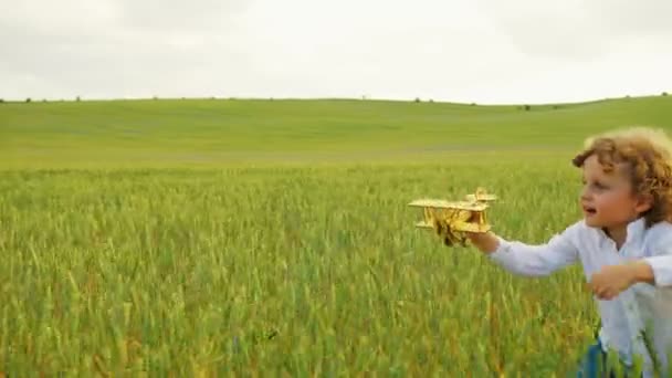 Glad liten pojke kör med gul trä flygplan leksak genom det gröna fältet. Stilig pojke leker med planet leksak på ängen. Närbild — Stockvideo