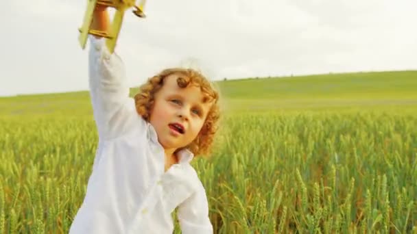 Niño feliz con rizos corriendo por el campo verde con juguete de avión de madera. Niño guapo jugando con el avión de juguete amarillo en el día soleado. De cerca. — Vídeo de stock