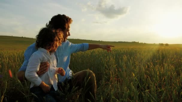 Feliz familia de padre e hijo sentado en el campo de trigo verde. Padre señalando con el dedo la hermosa puesta de sol. De cerca. — Vídeo de stock