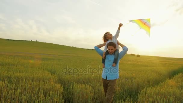 Jovem pai caminhando no campo de trigo verde com seu filho nos ombros. O miúdo a brincar com papagaios voadores. Pôr do sol — Vídeo de Stock