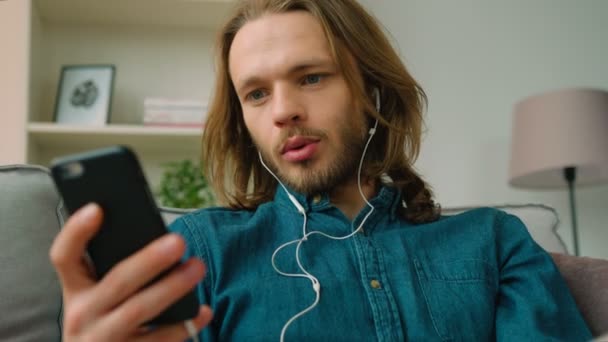 Молодой человек слушает музыку на своем смартфоне, сидя на диване в гостиной. Хипстер с длинными волосами. Закрыть — стоковое видео