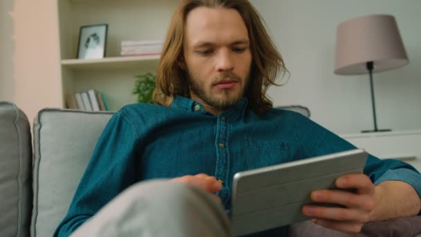 Jovem usando computador tablet enquanto sentado no sofá em casa. Hipster com cabelos longos navegando na internet e sorrindo — Vídeo de Stock