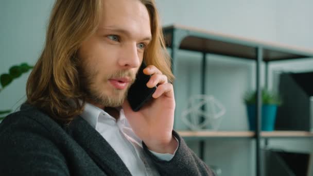 Молодой бизнесмен с длинными волосами разговаривает по телефону и работает за ноутбуком. Закрыть — стоковое видео