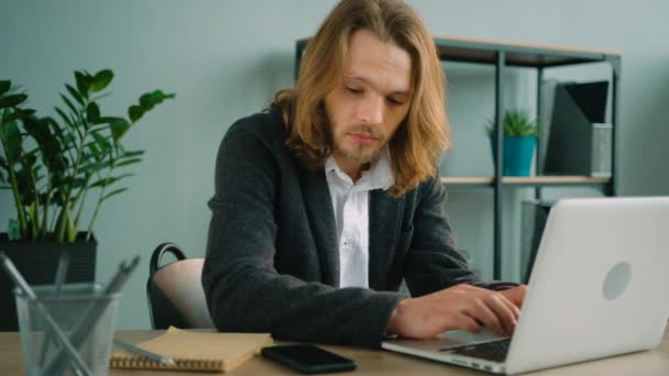 Ein junger Mann sitzt im Büro, telefoniert und tippt auf seinem Laptop. Mann mit langen Haaren schaut auf Bildschirm, checkt Informationen, während er mit dem Handy spricht. — Stockvideo