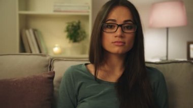 Oturma odasında kanepede oturan ve gülümseyerek kameraya bakarak çekici genç kadın portresi. Gece vakti