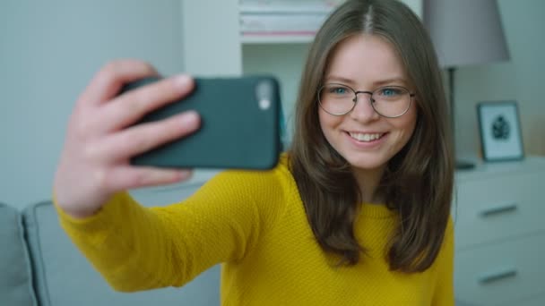 Hermosa chica adolescente en jersey amarillo haciendo selfies mientras está sentado en el sofá en la sala de estar. Chica sonriendo a la cámara y haciendo fotos. De cerca. — Vídeos de Stock