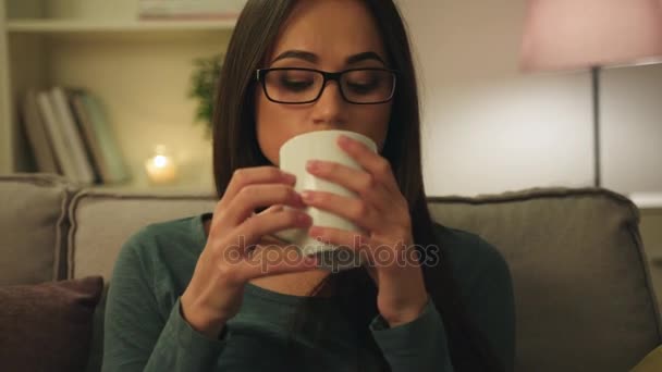 Aantrekkelijke jonge vrouw in glazen drinkng koffie, thee zittend op de Bank thuis op moment van de nacht. Kaars lampen — Stockvideo