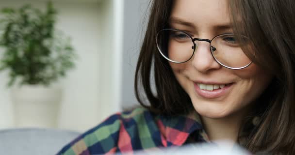 Adolescente menina lendo livro e sorrindo enquanto sentado no sofá na sala de estar. Fechar — Vídeo de Stock