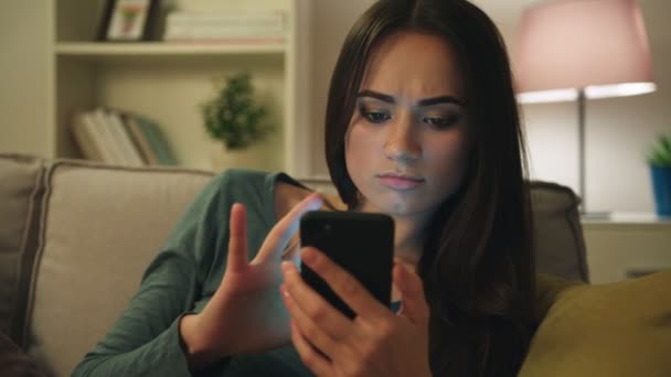 Aantrekkelijke jonge vrouw te typen op haar slimme telefoon, surfen op het internet terwijl zittend op de Bank in de woonkamer in de avond. Close-up — Stockvideo