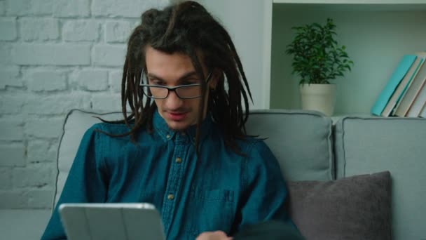 リビング ルームのソファに座って彼のタブレット デバイスでビデオ通話で感情的に話してドレッドヘアを持つ若い男. — ストック動画
