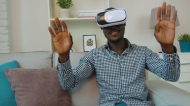 Oturma odasında kanepede, VR kulaklık kullanarak Afrika kökenli Amerikalı zenci. Aroung seyir ve sanal nesneleri dokunmadan çıktıktan adam