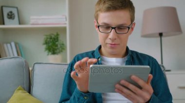 Tablet bilgisayar aygıtı kullanarak ve oturma odasında kanepede otururken gülümseyen yakışıklı genç öğrenci. Yakın çekim