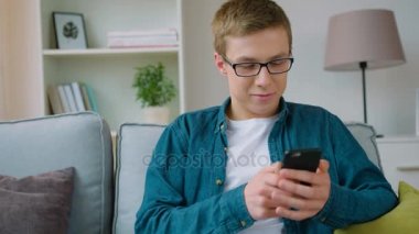 Oturma odasında kanepede oturan ve akıllı telefon kullanma yakışıklı genç öğrenci. Gülümseyen genç adam onun smartphone kullanarak