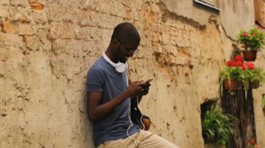 Gözlük ve T-shirt duvara yakın duran, akıllı telefon kullanarak, kameraya görünümlü ve şehir arka plan üzerinde elini sallayarak dostu Afro-Amerikan delikanlı. Açık.