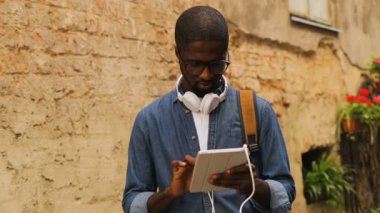 Şık genç Afro-Amerikan adam gözlüklü şehir duvarının yakınında duran arkadaşıyla Sohbet için tablet kullanma kulaklıklar ile.