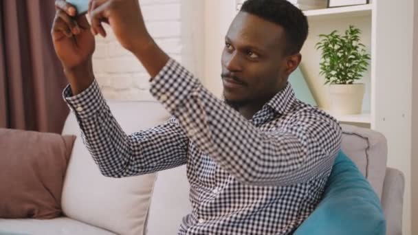 Negro africano americano hombre sentado en el sofá y hacer selfie en el teléfono inteligente cámara — Vídeo de stock