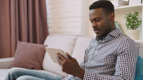 Negro afroamericano hombre sentado en el sofá y el uso de tableta dispositivo informático. Tocando en la pantalla táctil. De cerca. Vista lateral — Vídeo de stock