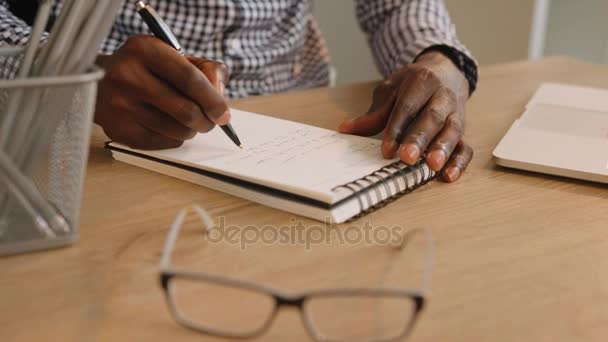 Чёрный африканский бизнесмен-американец пишет заметки в ежедневнике в офисе. Пишу идеи, планы. Закрыть — стоковое видео