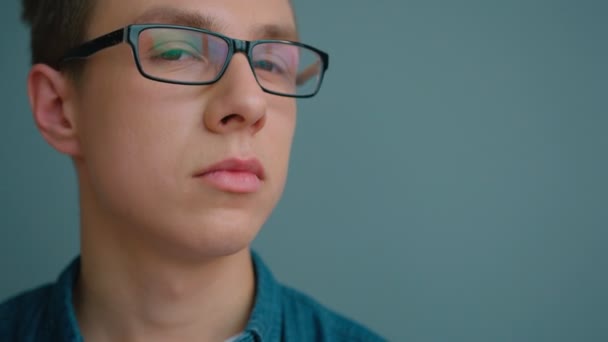 Retrato de estudante universitário em óculos sorrindo e olhando para a câmera no fundo da parede azul. Fechar — Vídeo de Stock