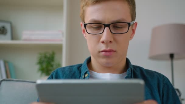 Przystojny, młody student za pomocą urządzenia komputera typu tablet, siedząc na kanapie w salonie. Z bliska — Wideo stockowe