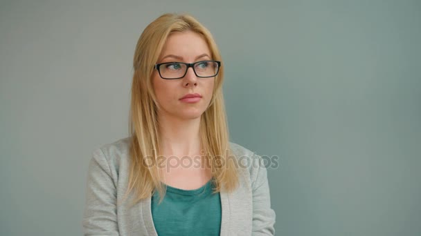 Vacker blond kvinna i glasögon titta rakt på kameran och leende på grå bakgrund. Porträtt skott — Stockvideo
