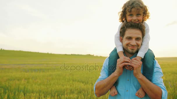 Portrét šťastný otec a syn sedí na ramenou. Otec a málo slunce s procházku v zelené pšeničné pole. Zblízka. Usmíval se na kameru — Stock video