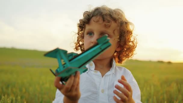 Κοντινό πλάνο πορτρέτο του ευτυχισμένο λίγο σγουρά αγόρι παίζει με μπλε ξύλινο αεροπλάνο παιχνίδι στο πεδίο πράσινο σιτάρι. Κοντινό πλάνο — Αρχείο Βίντεο