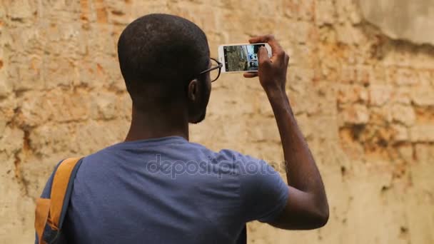 Achteraanzicht van jonge african american man in glazen en T-shirt in stad in de buurt van de muur staan, met behulp van slimme telefoon voor het maken van foto's. Buiten. — Stockvideo
