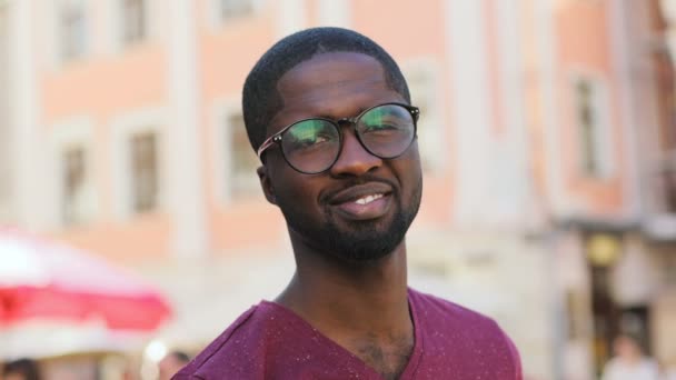 Retrato de jovem afro-americano de óculos e camiseta olhando para o lado, voltando-se para a câmera e sorrindo no fundo da cidade. Ao ar livre . — Vídeo de Stock