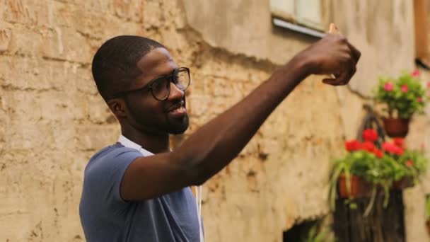 Atraente jovem afro-americano de óculos e camiseta em pé na cidade perto da parede, usando telefone inteligente para fazer vídeo. Ao ar livre . — Vídeo de Stock