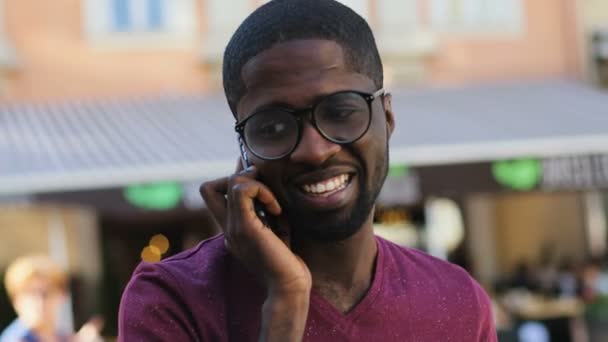 Портрет привлекательного молодого африканского мужчины в очках и футболке, разговаривающего по мобильному телефону с другом на фоне города . — стоковое видео