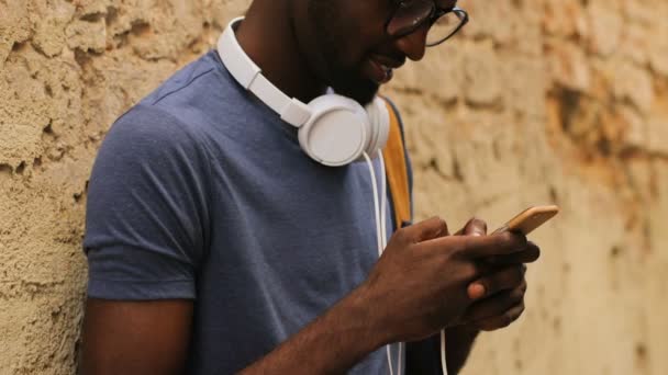 Porträtt av vänliga unga afroamerikanska man i glasögon och T-shirt står nära väggen och använder smarta telefonen mot staden bakgrund. Utomhus. — Stockvideo