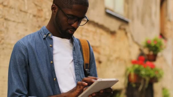 Vista lateral del retrato del joven afroamericano en gafas usando tableta para charlar con un amigo parado cerca de la pared en la ciudad . — Vídeos de Stock