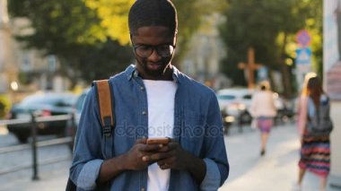 Genç Afrika turistik akıllı telefon kullanarak, kameraya bakıyor ve şehir merkezi ayakta ile gülümseyen mavi gömlekli. Açık.