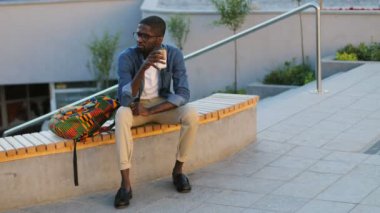 Merdiven zemin üzerine bankta otururken kahve içme kulaklıklarla bardaklarda çekici Afrika öğrenci. Açık.