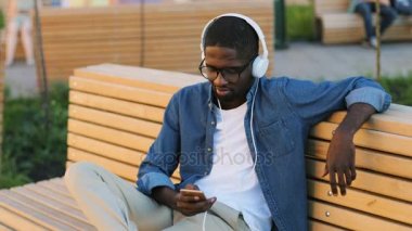 Yakışıklı genç Afrika adam akıllı telefon ve kulaklık kullanarak müzik City bankta siting için dinlemek için gözlük.