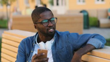 Şehirde bankta siting arkadaşıyla Sohbet için akıllı telefon kullanarak kulaklık ile gözlüklü genç Afrika adam portresi.