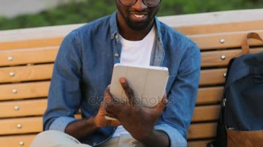 Güzel Afrika delikanlı gülen, parktaki bankta siting arkadaşıyla Sohbet için tablet kullanarak gözlük.